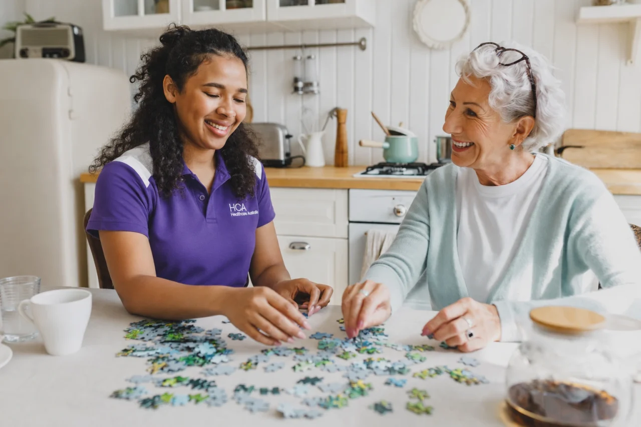 Eldery lady with support worker smiling doing a jugsaw puzzle on kitchen table